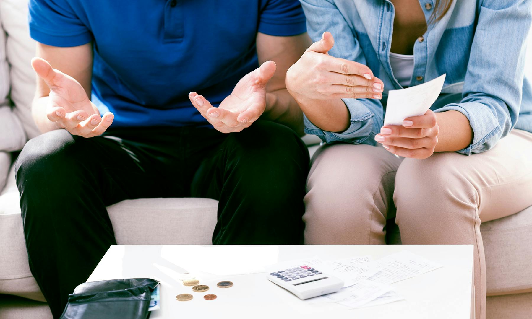 Photo of two people sitting on a couch reviewing bills and receipts, gesturing while talking, with a calculator, coins and a wallet on the coffee table in front of them